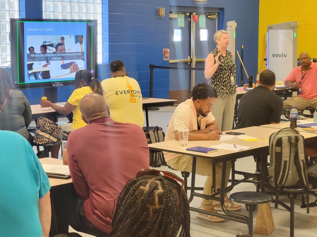 Leader instructing many people sitting at tables looking at a promethean board with a presentation on display